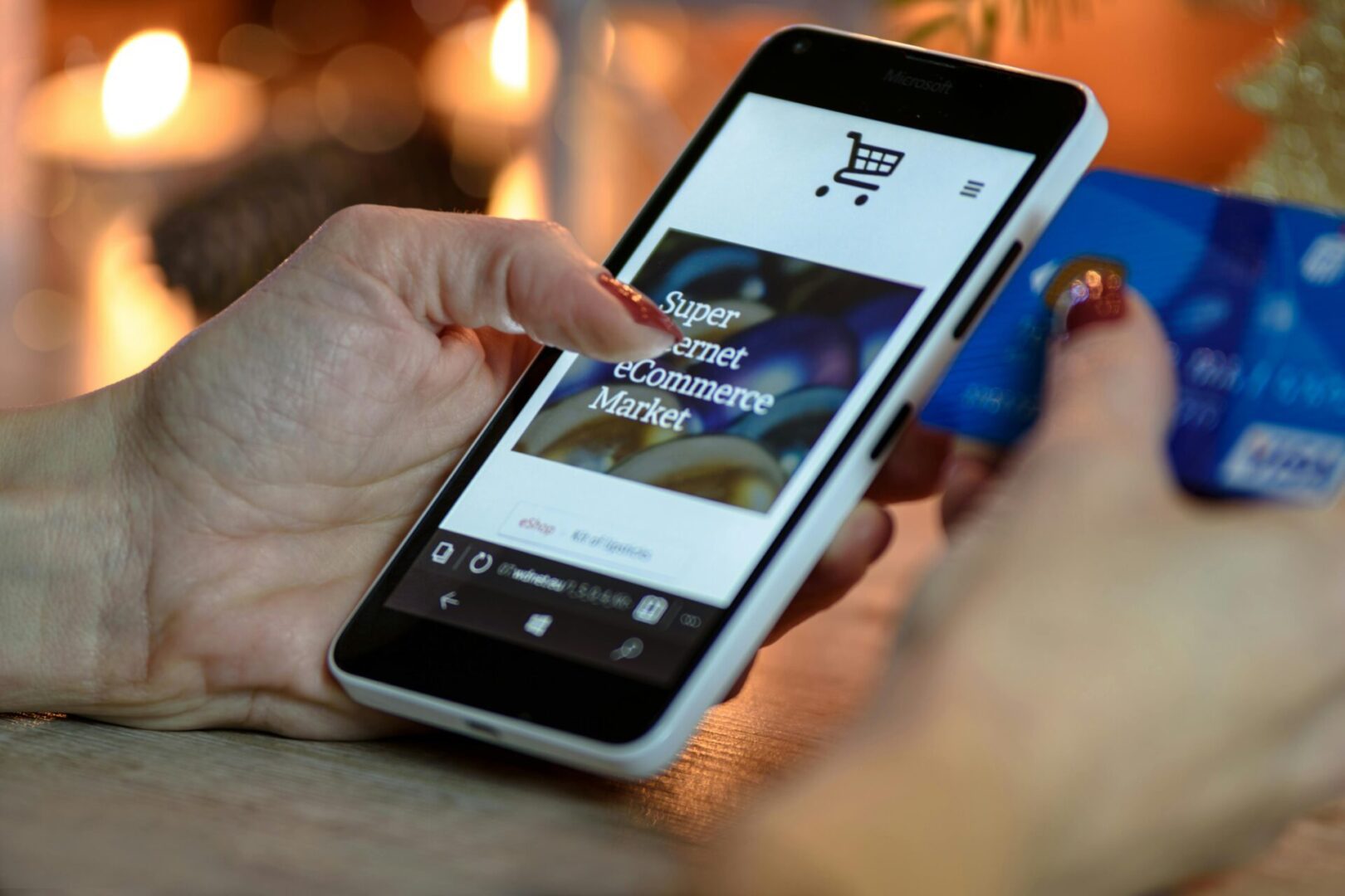 Close-up of a person's hands using a smartphone and a credit card for online shopping. The smartphone screen displays a webpage with the words "Super Internet eCommerce Market" visible. A shopping cart icon is also present on the screen. A credit card, light blue with gold accents, is held alongside the phone. The person's hand, holding the phone with the thumb positioned to interact with the screen, has a hint of reddish-brown nail polish. The background is out of focus, with a blurred image of warm-toned candles in the background. The image is taken at a low angle looking slightly up at the hands.