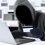 A faceless figure in a hoodie and gloves holds a credit card in front of a laptop, suggesting cybercrime in a room with blurred computer equipment.